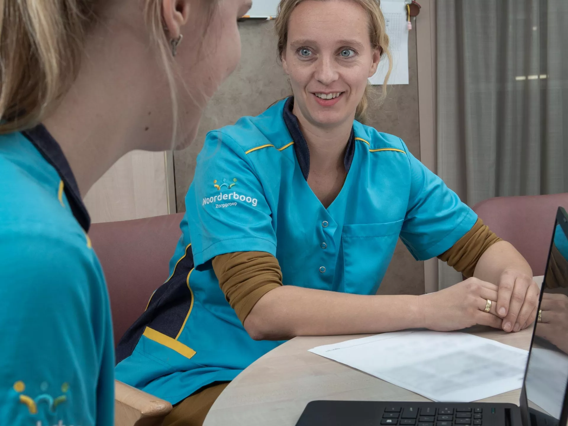 Twee zorgprofessionals in gesprek aan een tafel.