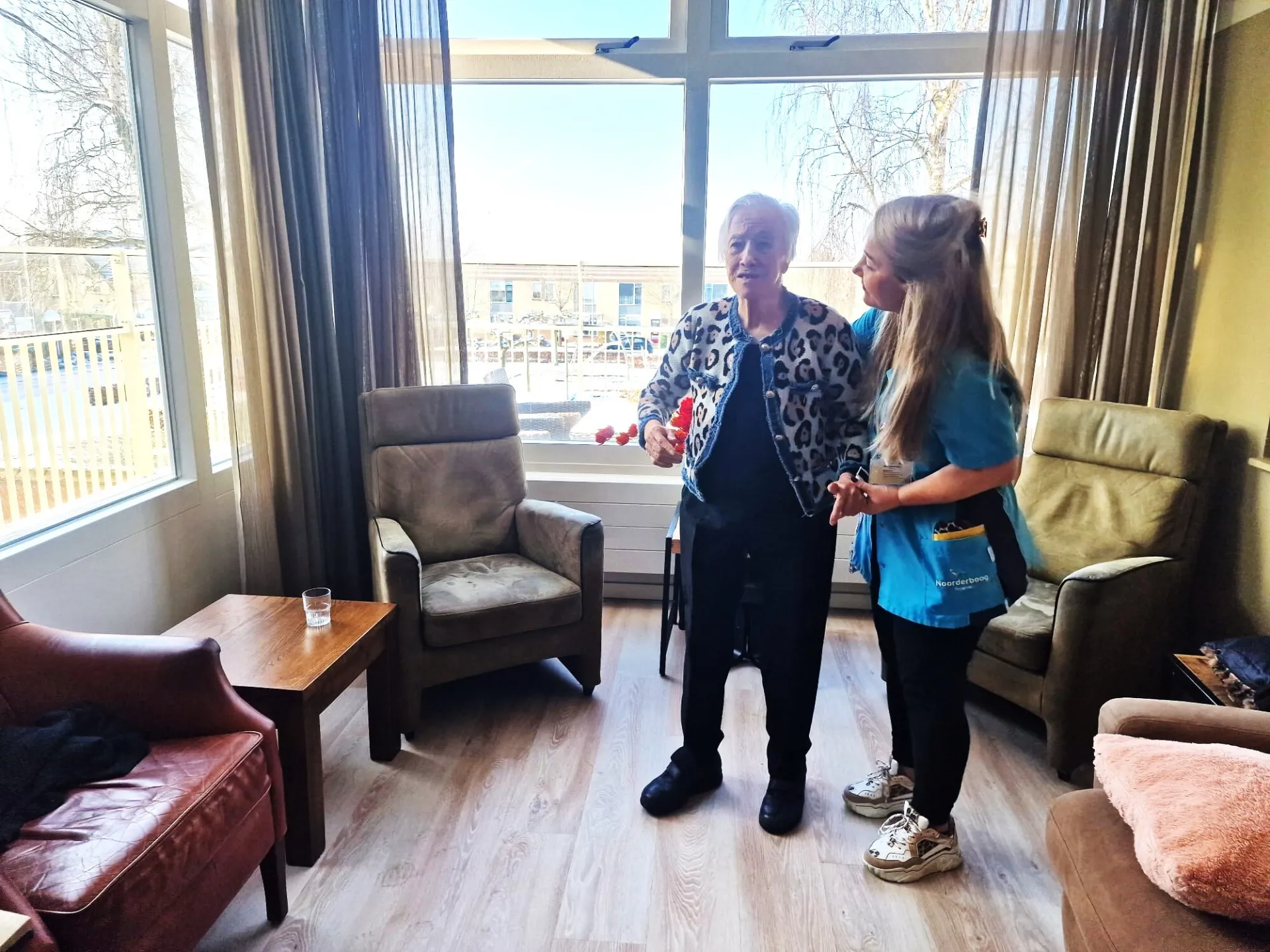 Verzorgende en bewoner in de woonkamer bij een groot raam.
